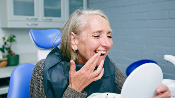 A cheerful mature woman checks her smile in a hand mirror during a dental appointment, sitting in the dental chair. The image conveys themes of health, care, and confidence in dental treatment.